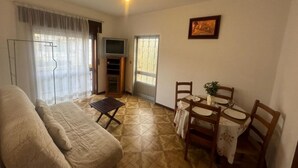 A living room with a sofa, a small table with chairs, a television, and a door leading to another room. - Appartement F2 Avec Jardin Cloture ET Fleurie (Medelo)