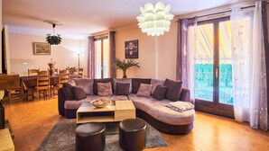 Un salon comprenant un canapé d’angle, une table basse, un coin repas avec des chaises et une table, ainsi qu’un balcon avec des rideaux.