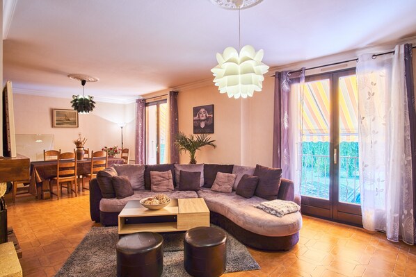Ein Wohnzimmer mit Ecksofa, Couchtisch, Essbereich mit Stühlen und Tisch sowie Balkon mit Vorhängen.