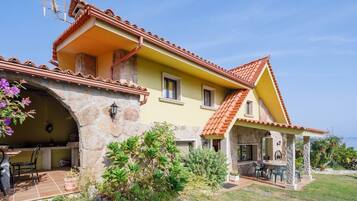 A two-story house with a stone archway, a patio with outdoor furniture, and a view of the sea.