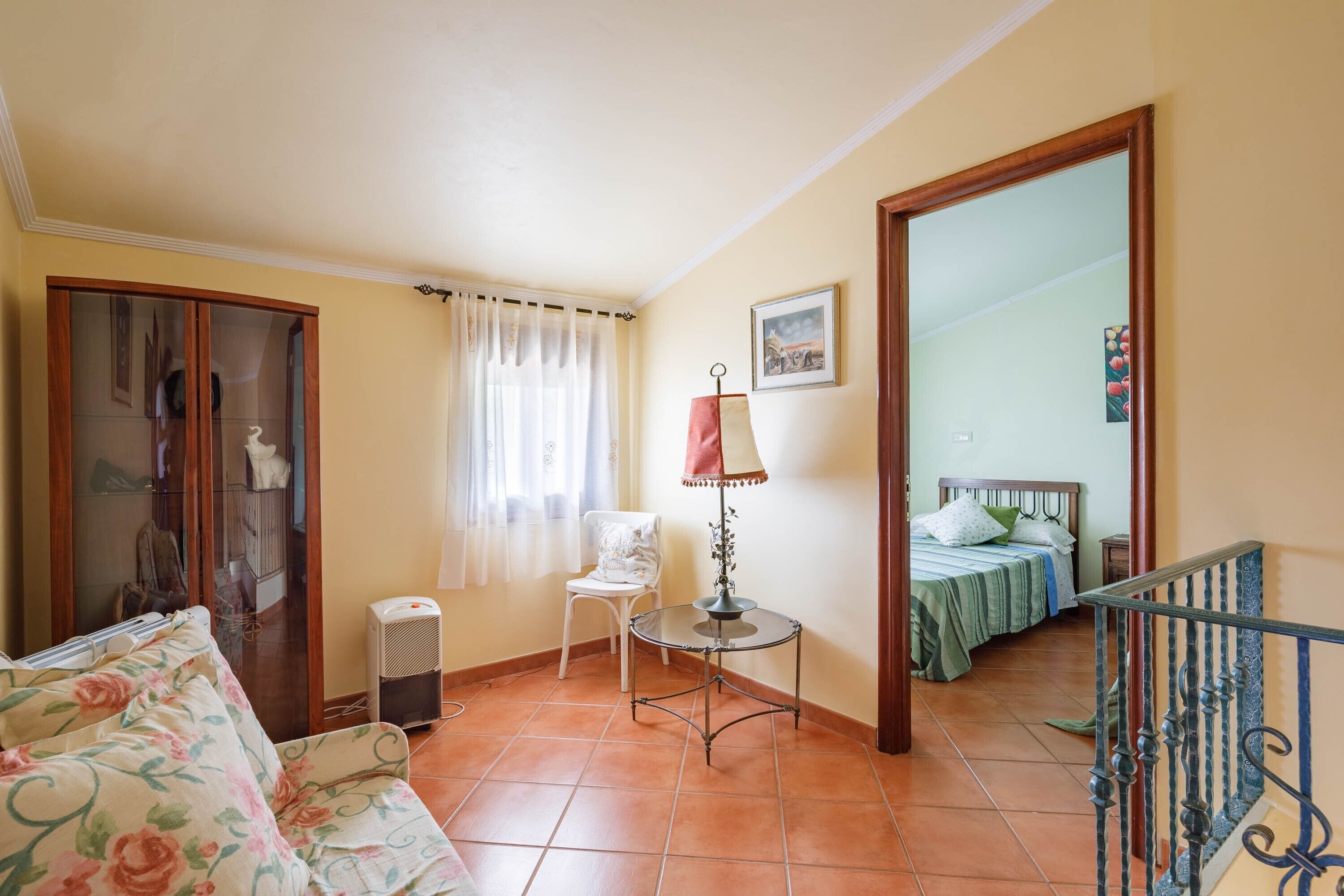 Une pièce comprenant un canapé, un fauteuil, une table avec une lampe, un lit recouvert d’une couverture verte et un grand miroir.