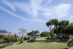 Un espace extérieur verdoyant, avec une table et des chaises, entouré d’arbres et offrant une vue sur la mer.