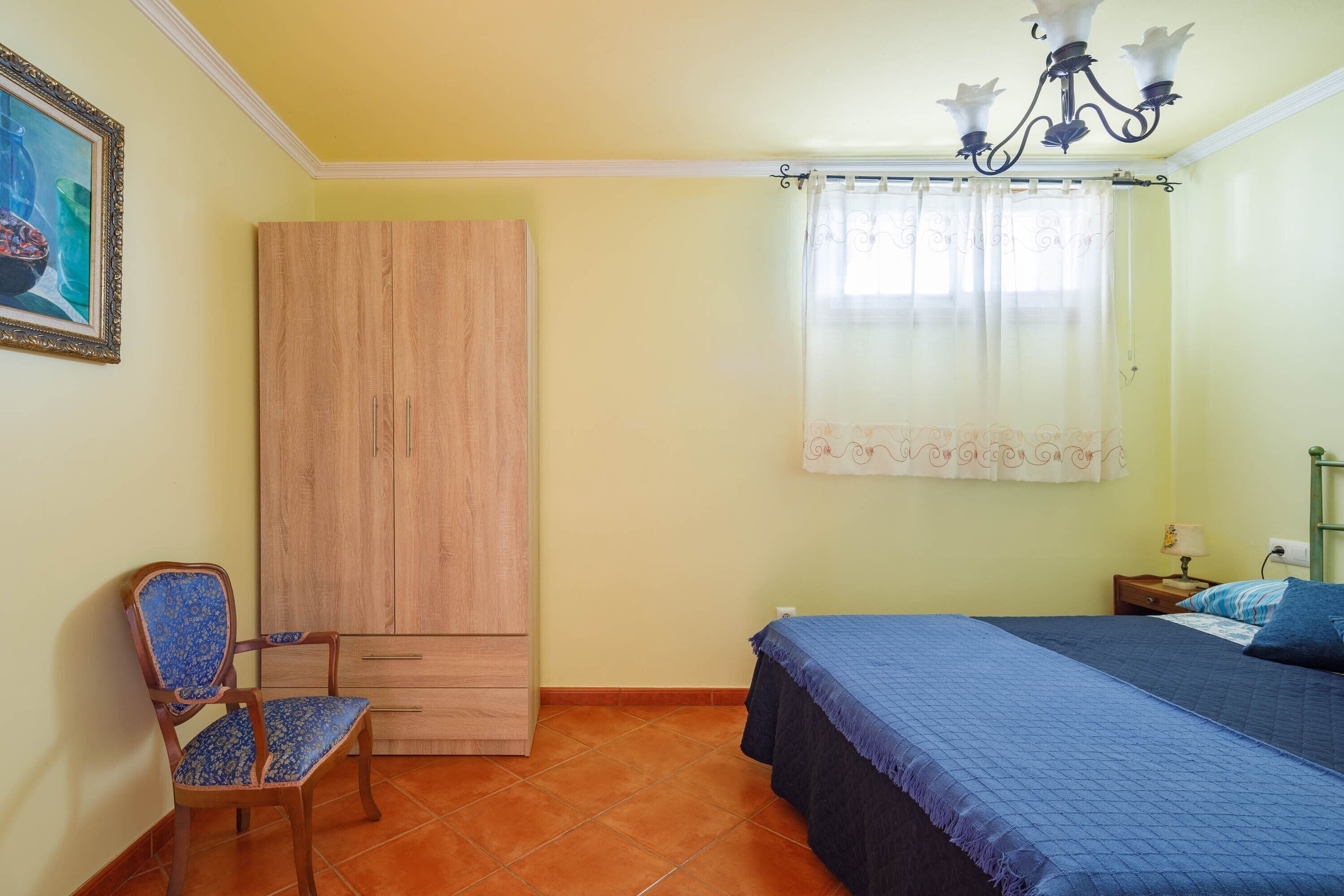 Un dormitori amb llit, una cadira, un armari, una finestra amb cortines i un penjador.