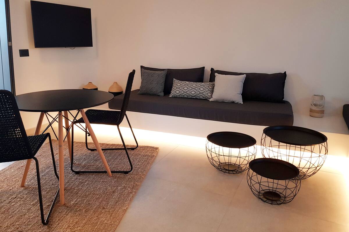 Ein modernes Wohnzimmer mit einer Couch, zwei schwarzen Sesseln, einem runden Tisch und drei Beistelltischen mit Drahtgestell.