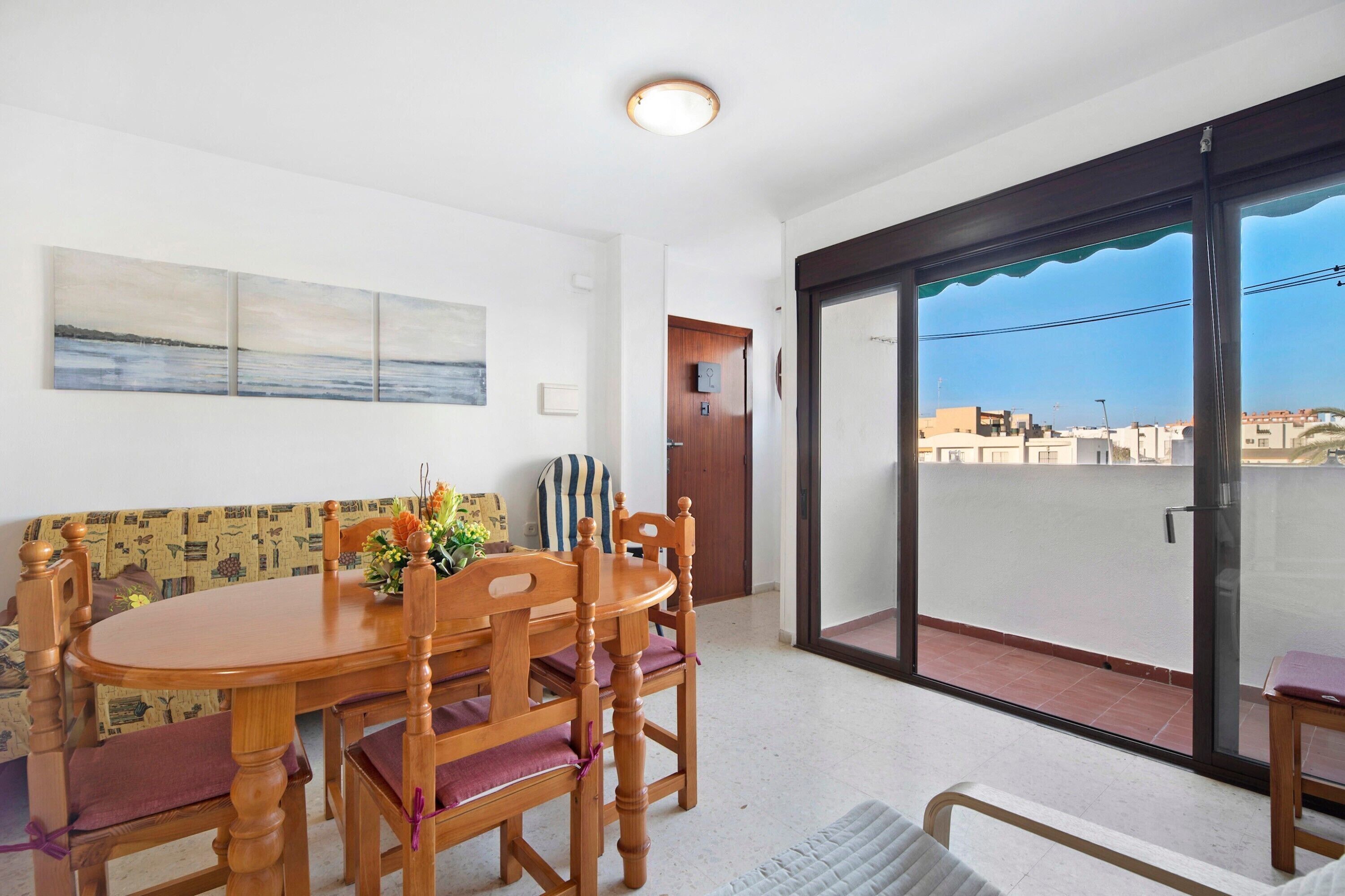 A dining area with a wooden table and chairs, a sofa, and a view of the city through a sliding glass door.