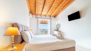 Ein Schlafzimmer mit Holzbalkendecke, einem Bett mit weißer Bettwäsche, einer Nachttischlampe, einem an der Wand befestigten Fernseher und einem Fenster mit Blick.