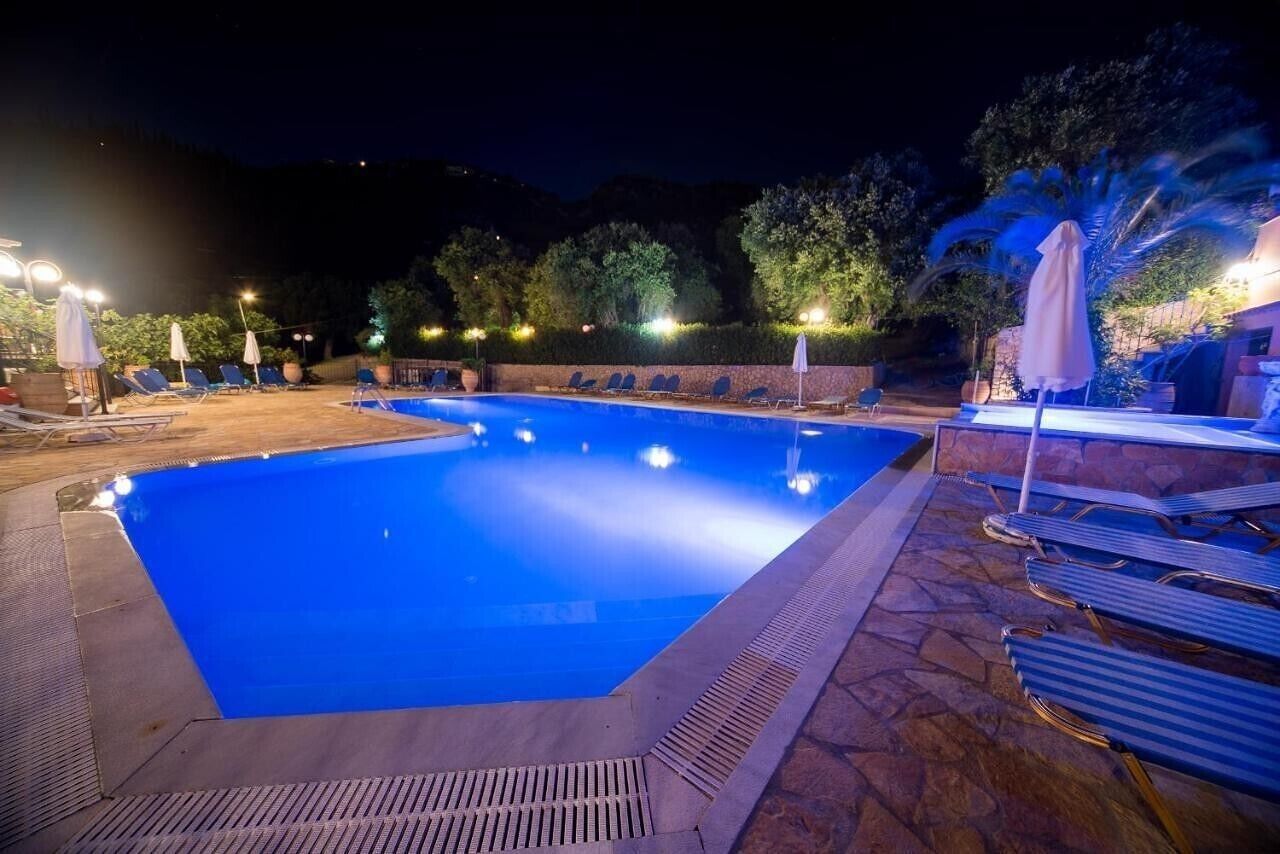 A nighttime swimming pool with lounge chairs and umbrellas, surrounded by stone walls and trees.