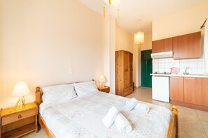Un dormitorio con cama, mesita de noche, lámpara y una zona de cocina con armarios y un refrigerador.