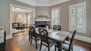 A dining area with a wooden table and chairs, a fireplace, and large windows with shutters.