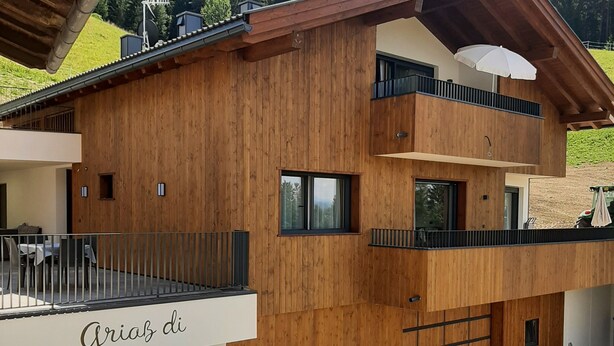 Een houten gebouw met twee verdiepingen, een balkon en een bord waarop 'Griaz di' staat, met uitzicht op een beboste heuvel.