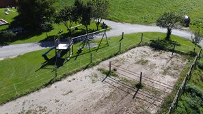 Vue aérienne d’une zone délimitée par une clôture, comprenant une aire de jeux, un chemin de terre et un champ d’herbe.
