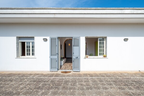 Un bâtiment à la façade blanche, doté de deux fenêtres avec volets et d’une porte d’entrée centrale qui mène à l’intérieur.