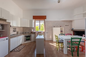 A modern kitchen with white cabinets, a central island, a dining table with colorful chairs, and a view through a window.