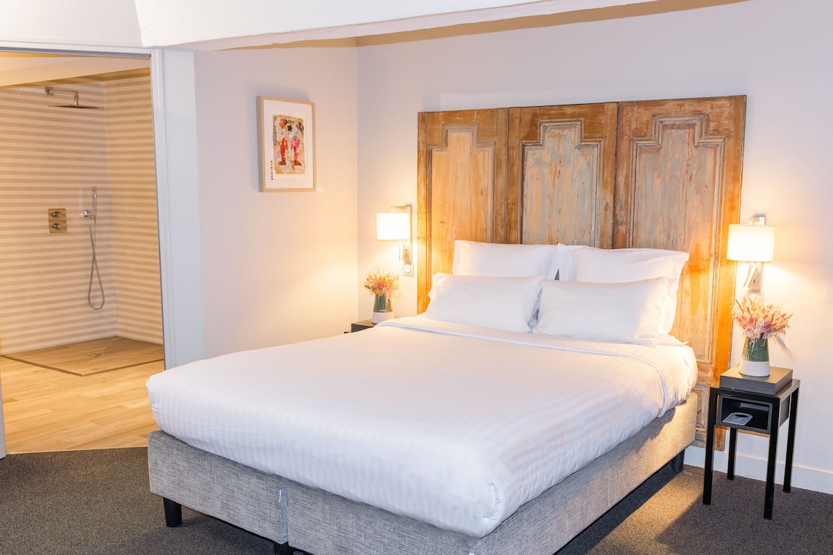 A neatly made bed with white linens, a wooden headboard, two bedside lamps, and a small bedside table with a vase of flowers.