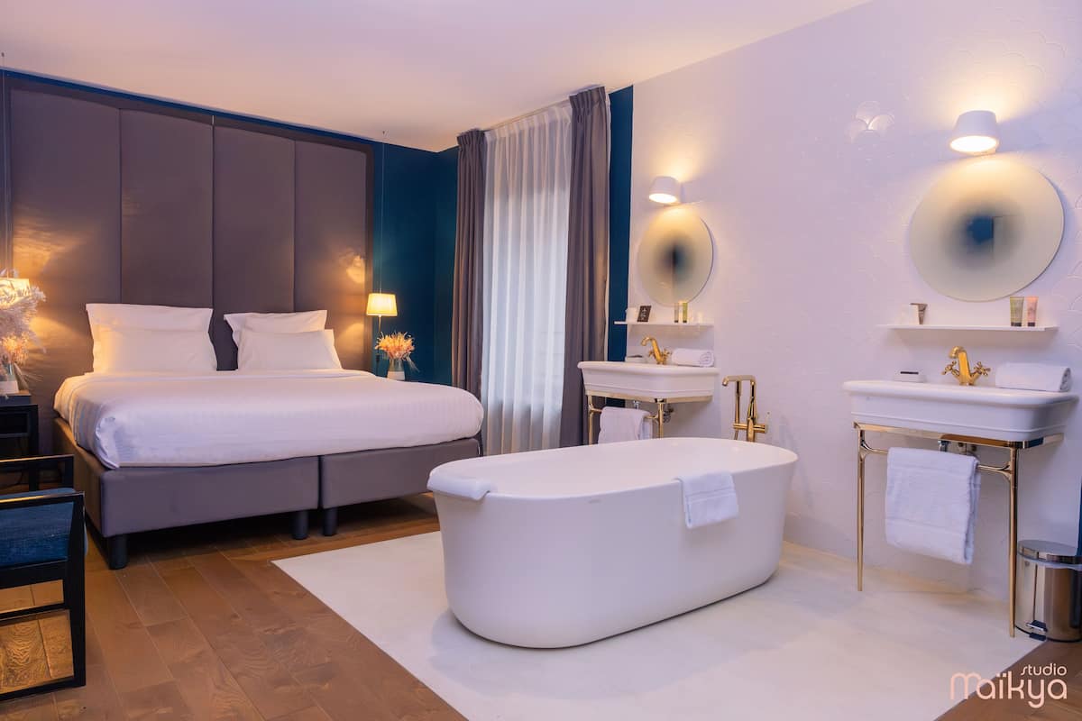 A modern hotel room with a large bed, a separate sitting area, a bathtub, a vanity with a mirror, and a small shelf with toiletries.
