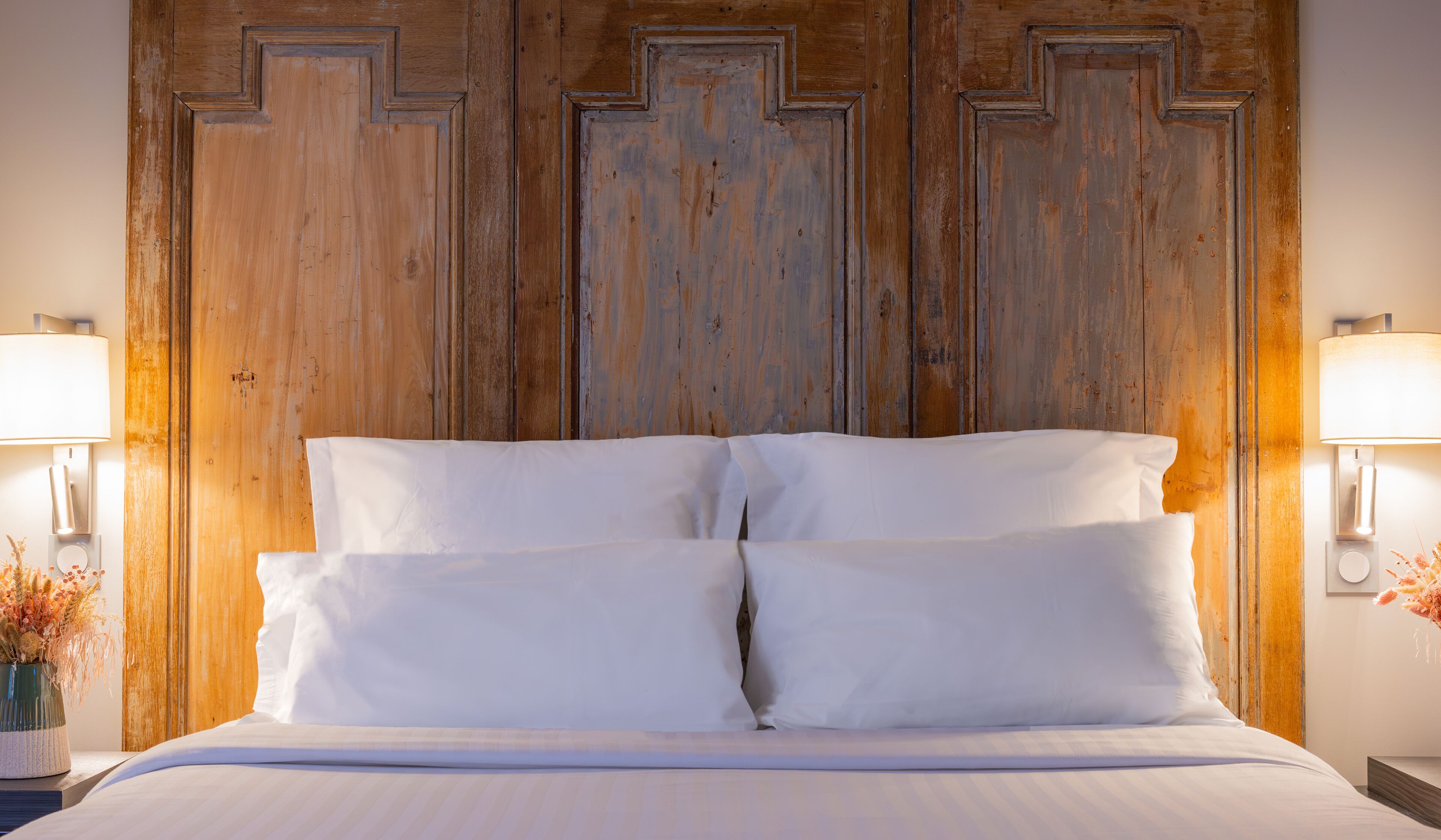 Un lit avec du linge de lit blanc et deux lampes de chevet, placé devant une tête de lit en bois ornée d’un motif géométrique.