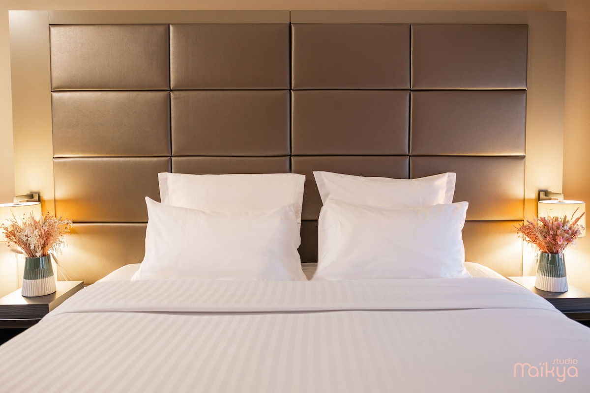 A hotel room with a large bed, padded headboard, white linens, and bedside lamps with floral arrangements.