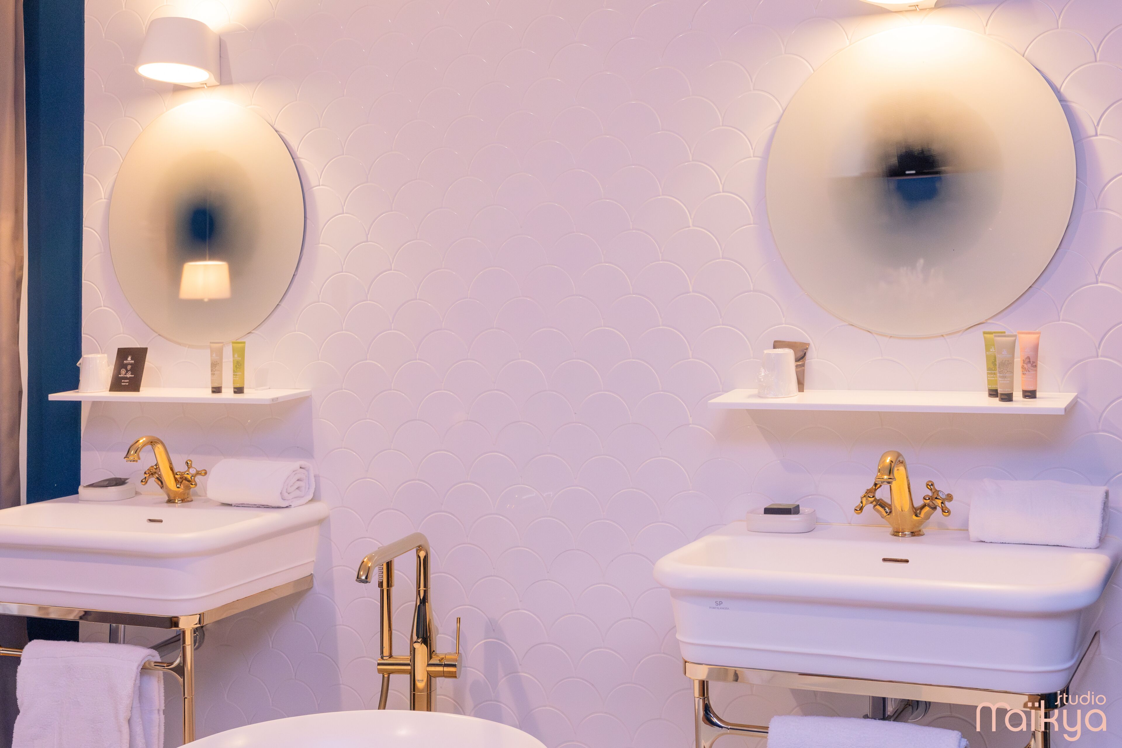 Baño con dos lavamanos, cada uno con grifo dorado y espejo encima. Hay repisas sobre los lavamanos con artículos de aseo.