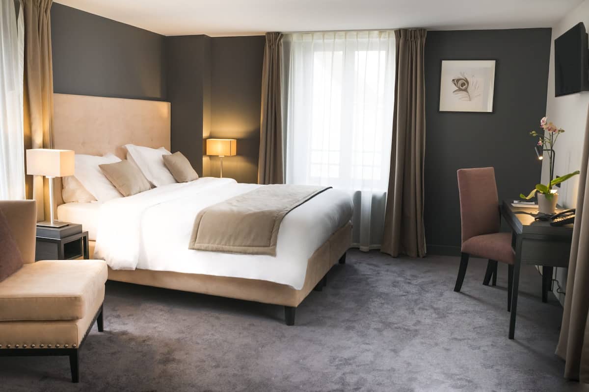 A hotel room with a large bed, a chair, a desk with a lamp, and a potted plant.