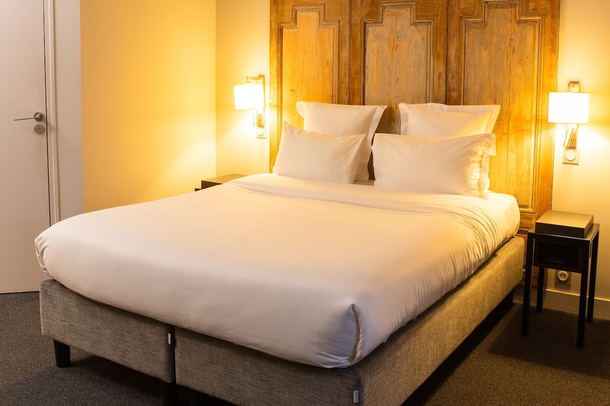 A bed with white linens, a wooden headboard, two bedside lamps, and a carpeted floor.