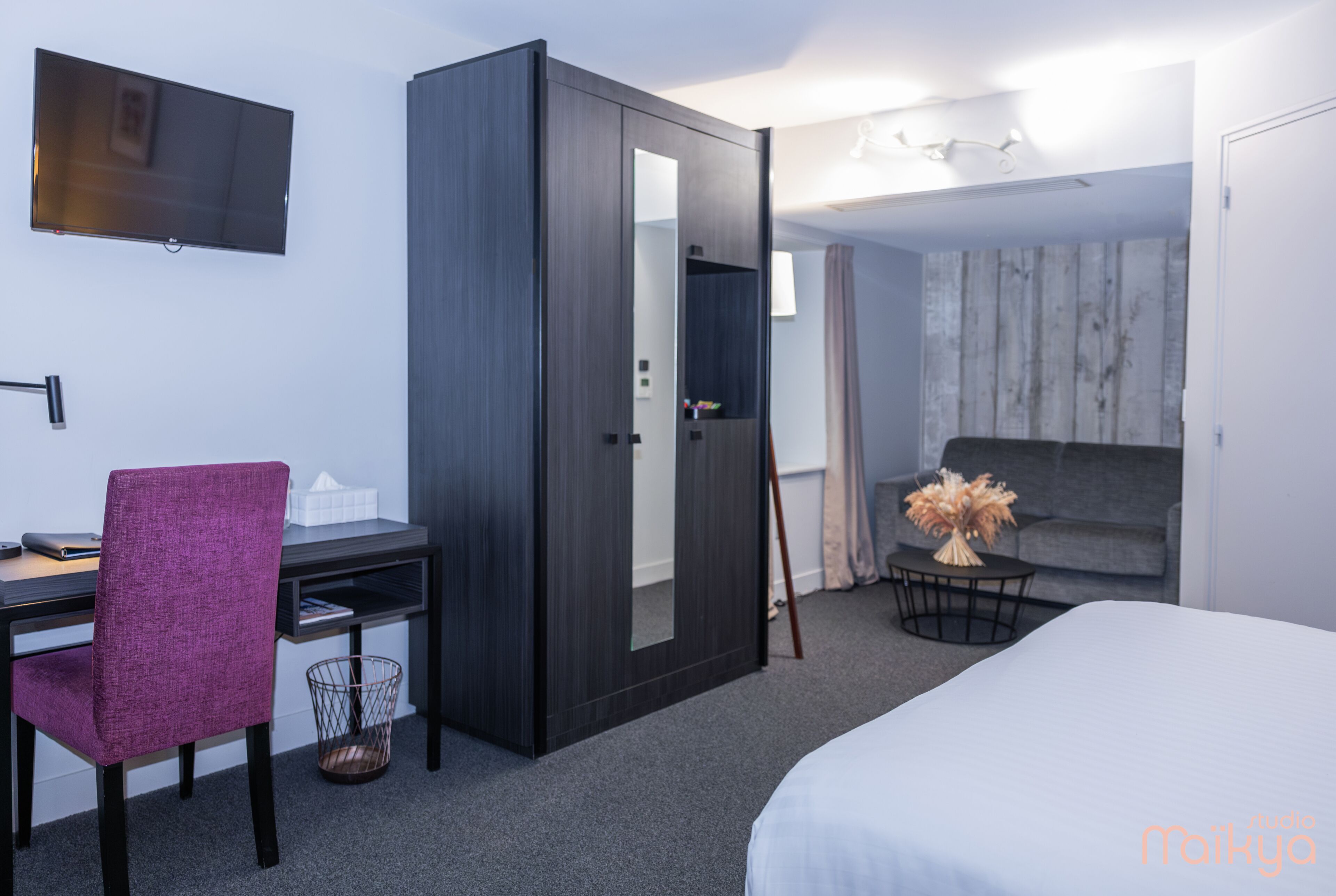 Habitación de hotel con cama, escritorio y silla, armario, televisor y una pequeña mesa con un florero.
