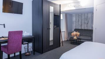 Habitación de hotel con cama, escritorio y silla, armario, televisor y una pequeña mesa con un florero.