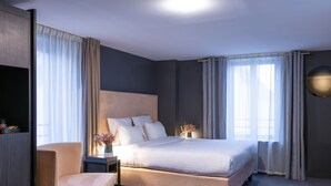 Una habitación de hotel moderna con una cama grande, una mesita de noche con un florero, una silla y un ventanal con cortinas.