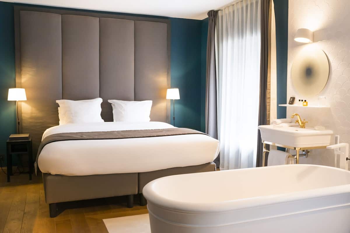A hotel room with a large bed, bedside lamps, a bathroom with a sink and mirror, and a bathtub.