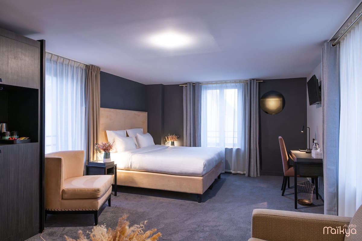 A modern hotel room with a large bed, a sitting area with a chair and a small table, and a desk with a chair.