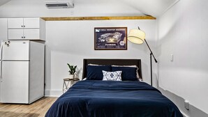 Un dormitorio con cama, mesita de noche, lámpara, un cartel y un refrigerador blanco.