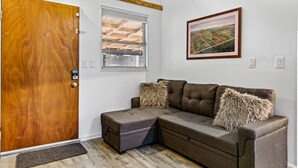 Una habitación con puerta de madera, ventana con persianas, sofá marrón con dos almohadas mullidas y un cuadro de paisaje enmarcado en la pared.