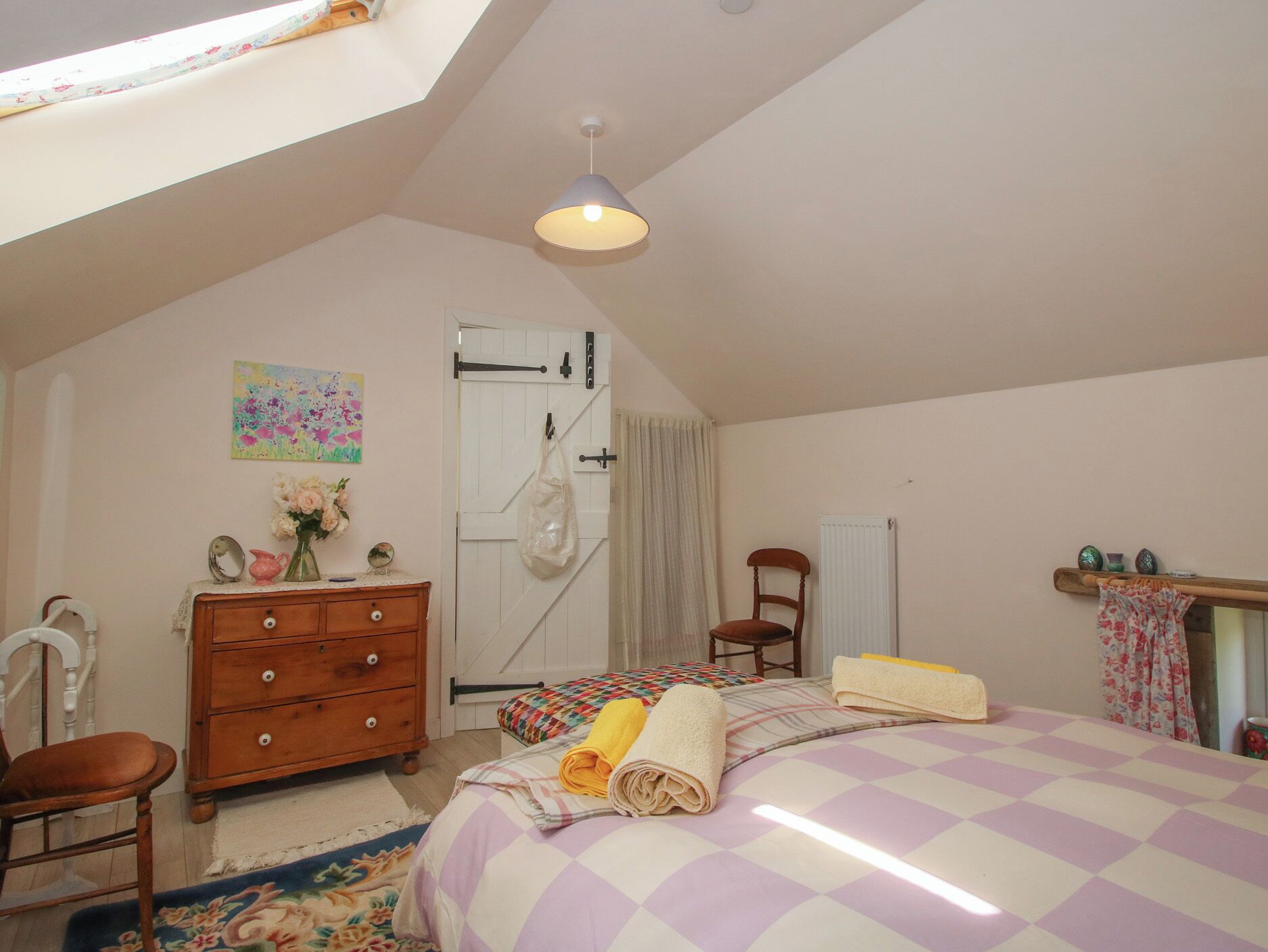 Una camera da letto con soffitto spiovente, un letto con copripiumino a quadri, una cassettiera in legno, una sedia, un termosifone e una finestra con tende.