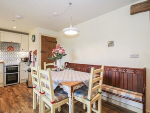 Un comedor con mesa de madera, sillas, un banco, un jarrón de flores y una cocina al fondo.