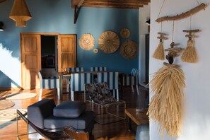 Uma sala de estar moderna com área de jantar, apresentando um sofá, uma mesa de centro e uma mesa de jantar com cadeiras listradas. Há também adornos na parede e uma peça de macramé pendente.