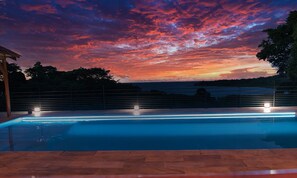 Piscina com vista para o pôr do sol, cercada por uma grade e árvores.