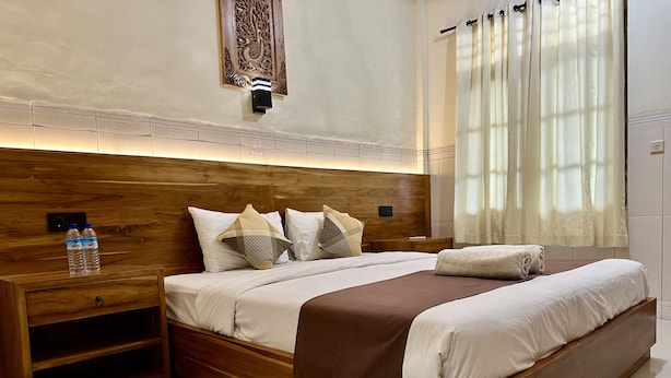 A modern bedroom with a wooden headboard, a bedside table with a water bottle, a large bed with a brown throw blanket, and a decorative wall piece above the bed. - GILI BEACH WIND (Gili Trawangan)