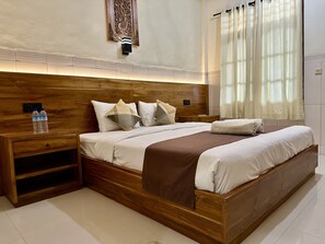 A modern bedroom with a wooden headboard, a bedside table with a water bottle, a large bed with a brown throw blanket, and a decorative wall piece above the bed. - GILI BEACH WIND (Gili Trawangan)