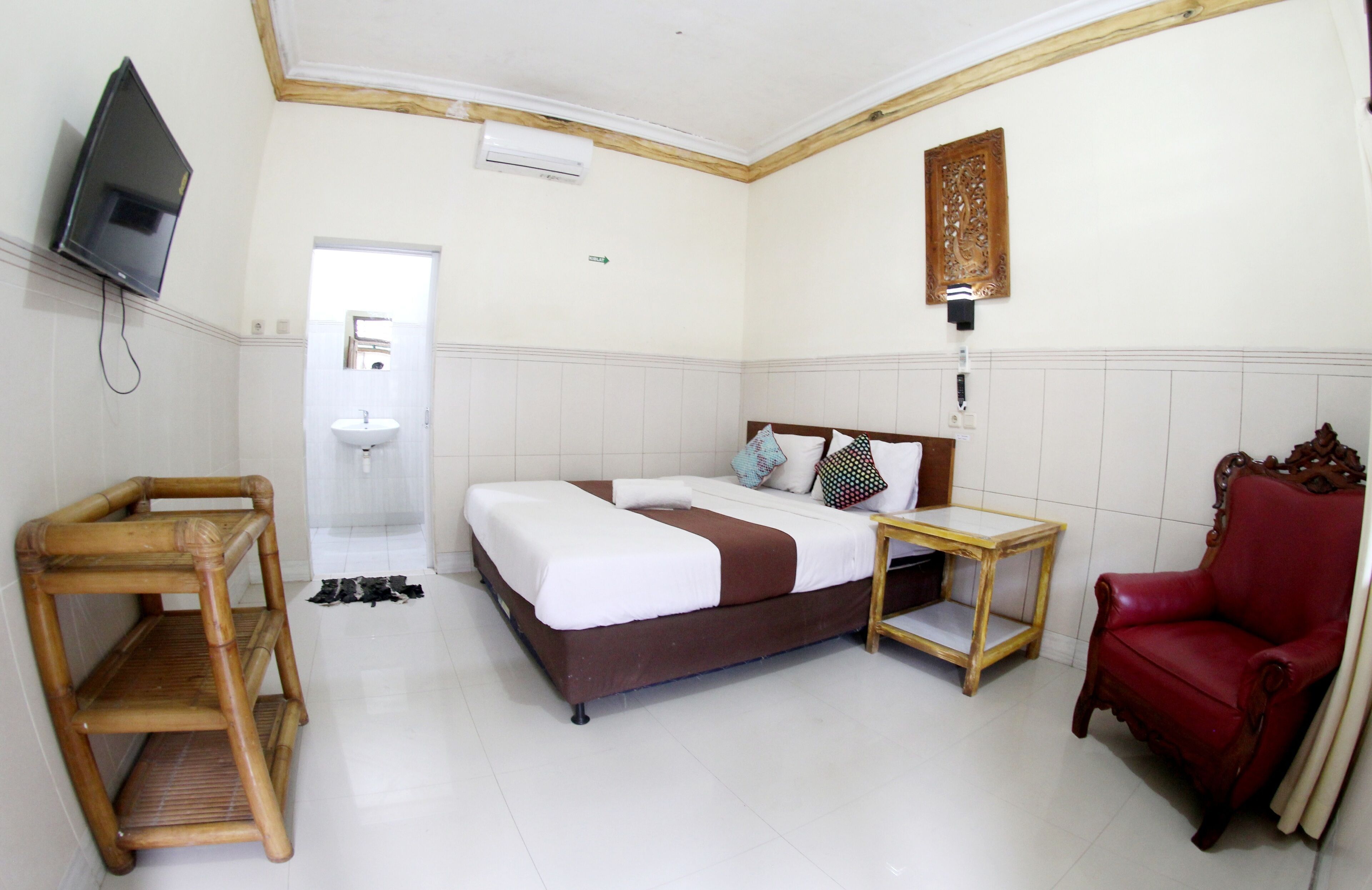 A hotel room with a bed, a red armchair, a wooden side table, a television, and a bathroom visible through an open door.