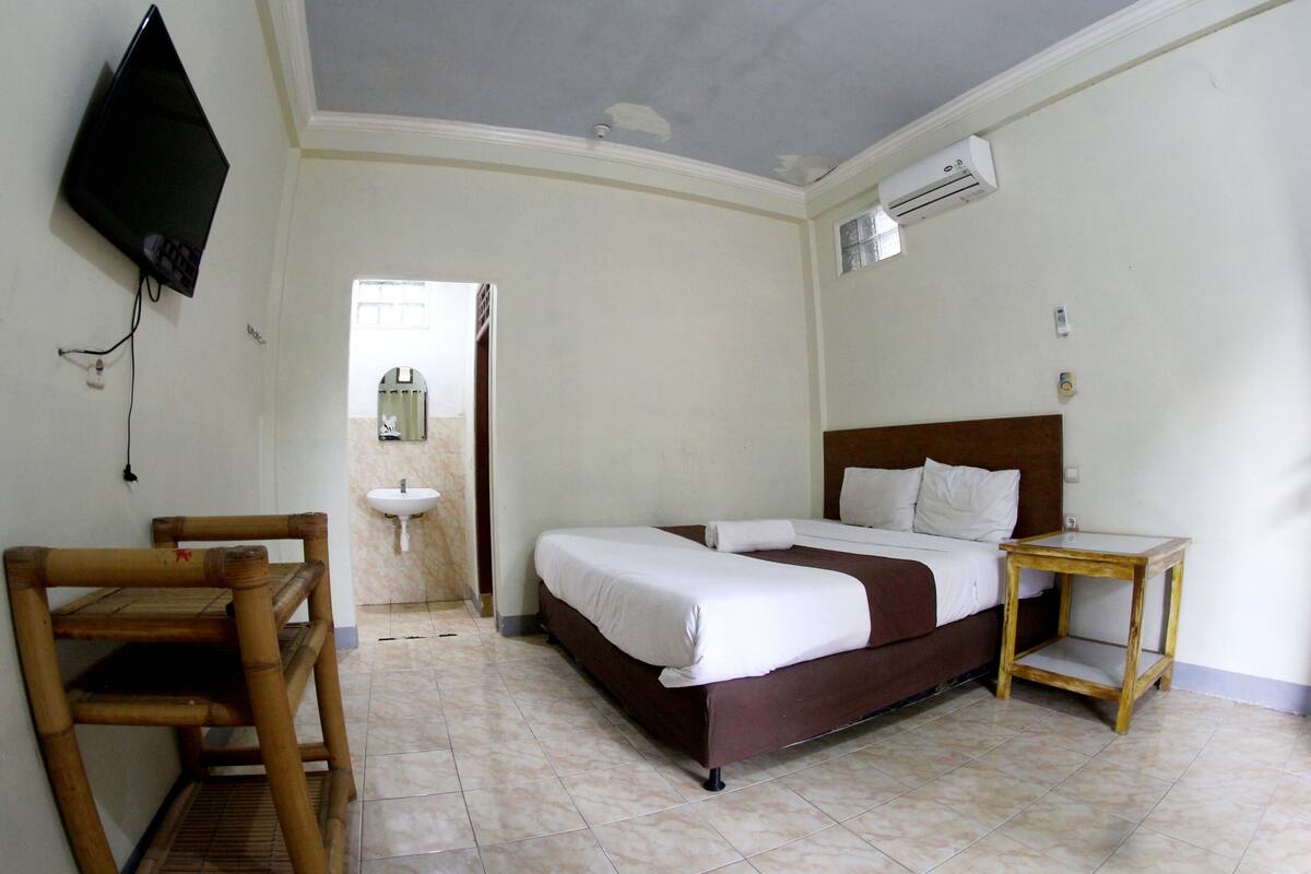 Ein Hotelzimmer mit einem Bett, einem Holzstuhl, einem kleinen Tisch, einem an der Wand befestigten Fernseher sowie einem Badezimmer mit Waschbecken und Spiegel.