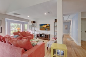 A spacious living room with a large window, a red sofa, a wooden coffee table, and a painting of a sunset.