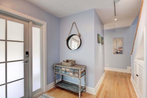 A hallway with a wooden floor, a door with frosted glass panels, a vintage-style console table with a woven basket, and a wall-mounted mirror.