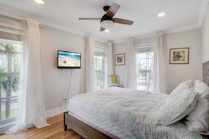 A bedroom with a bed, a ceiling fan, a television, and large windows with curtains.