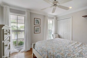 Un dormitorio con cama, ventilador de techo, dos ventanas con cortinas y una puerta.