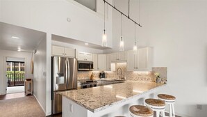Een moderne keuken met witte kasten, roestvrijstalen apparatuur, een groot eiland met barkrukken, hanglampen en uitzicht op een balkon.