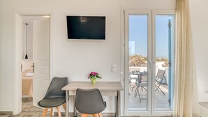 Een kamer met een aan de muur bevestigde televisie, een tafel met twee stoelen, een potplant en een glazen schuifdeur die leidt naar een buitenruimte.
