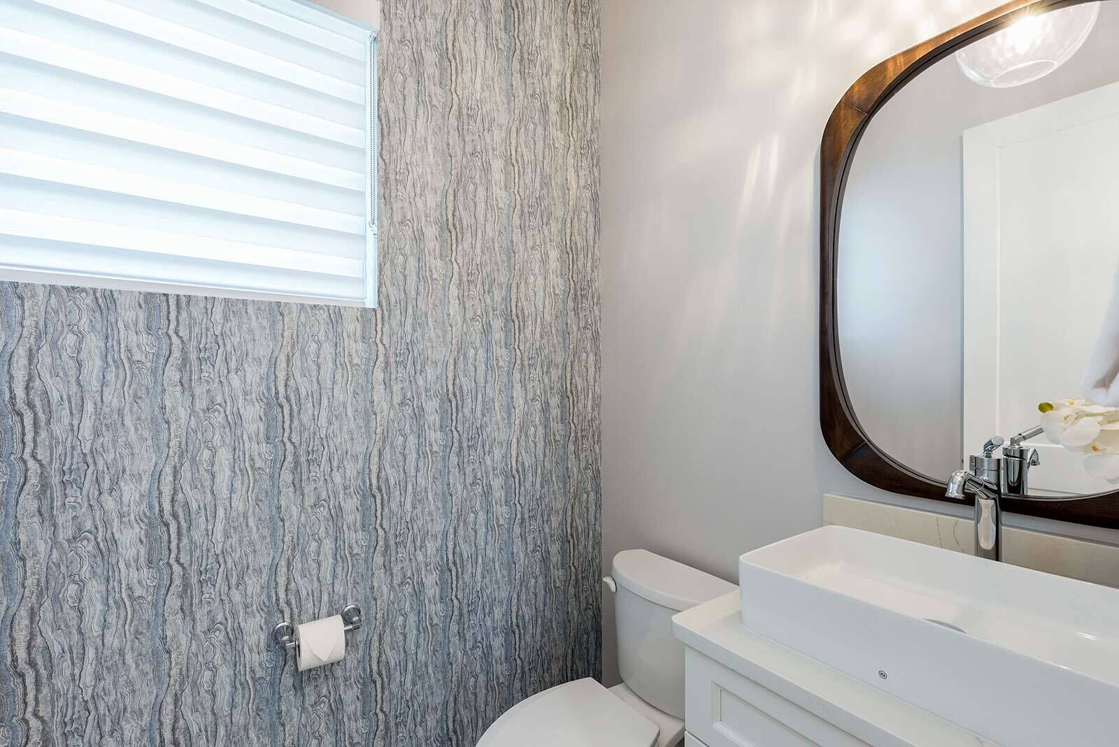 Un baño con pared de mármol, un inodoro, un lavabo con espejo y una ventana con persianas.