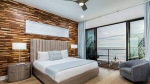 A modern bedroom with a large bed, a ceiling fan, a view of the sea, and a balcony with a chair.