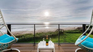 A balcony with wicker chairs and a table, overlooking a beach with a wooden deck and a fence.