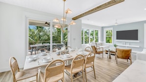 A modern dining area with a large table set for six, surrounded by wicker chairs, a chandelier, and large windows offering a view of palm trees.