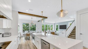A modern kitchen with white cabinetry, a marble island, and a dining area with a view of greenery.